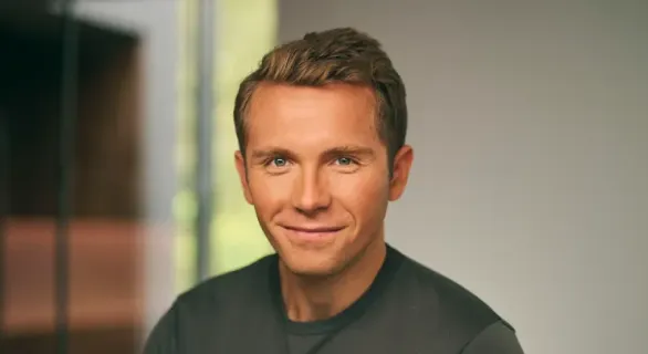 Smiling man with short light brown hair wearing a dark shirt, with a blurred background.